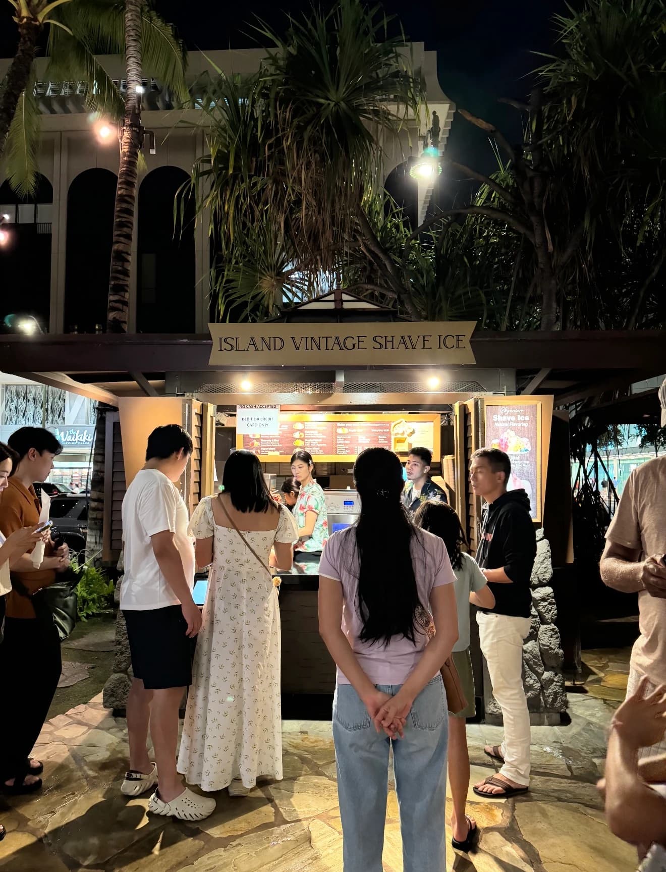 Late-night crowd ordering at a tropical shave-ice kiosk beneath palm trees