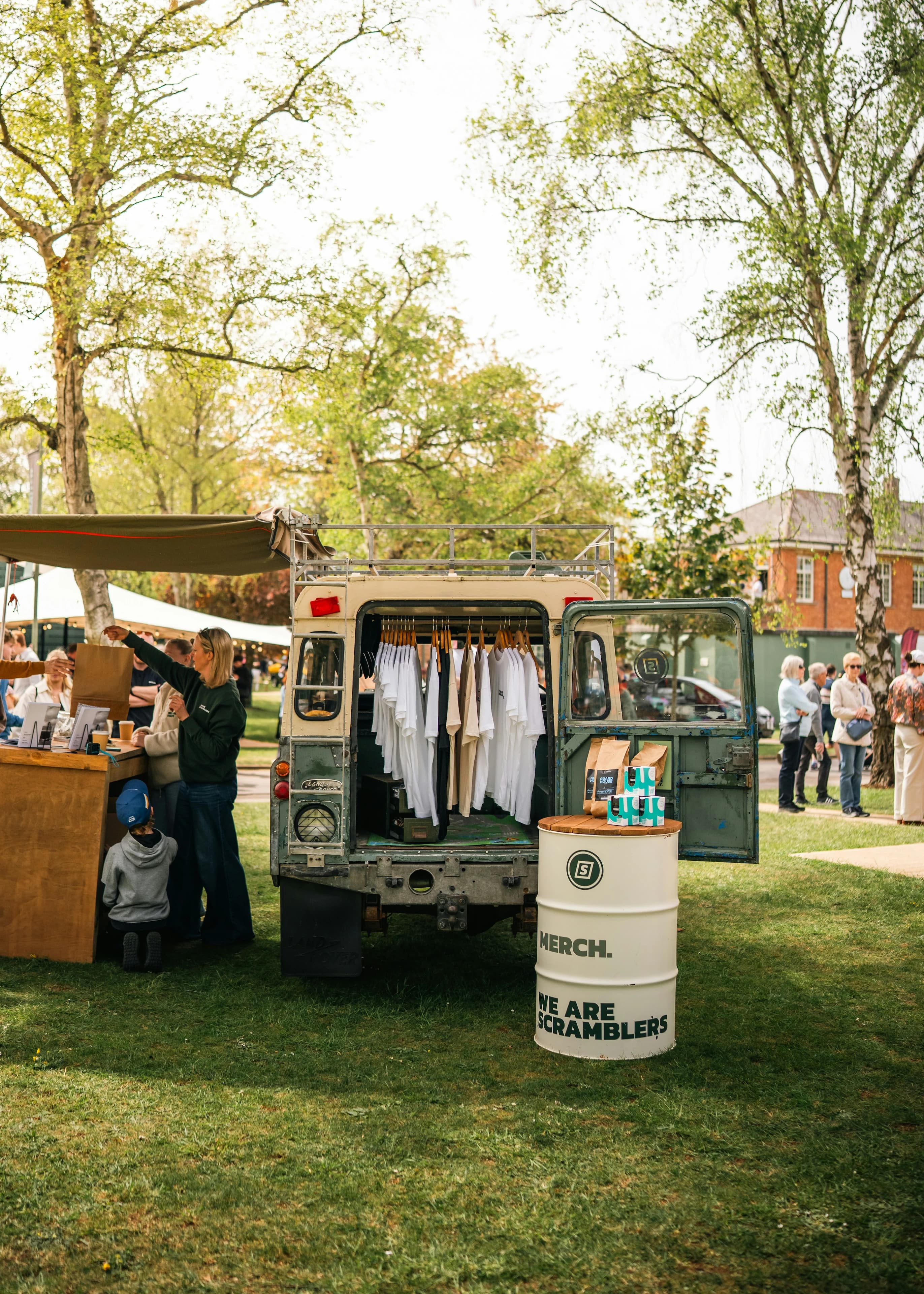 Open-air market stall selling t-shirts from a vintage Land Rover under the trees